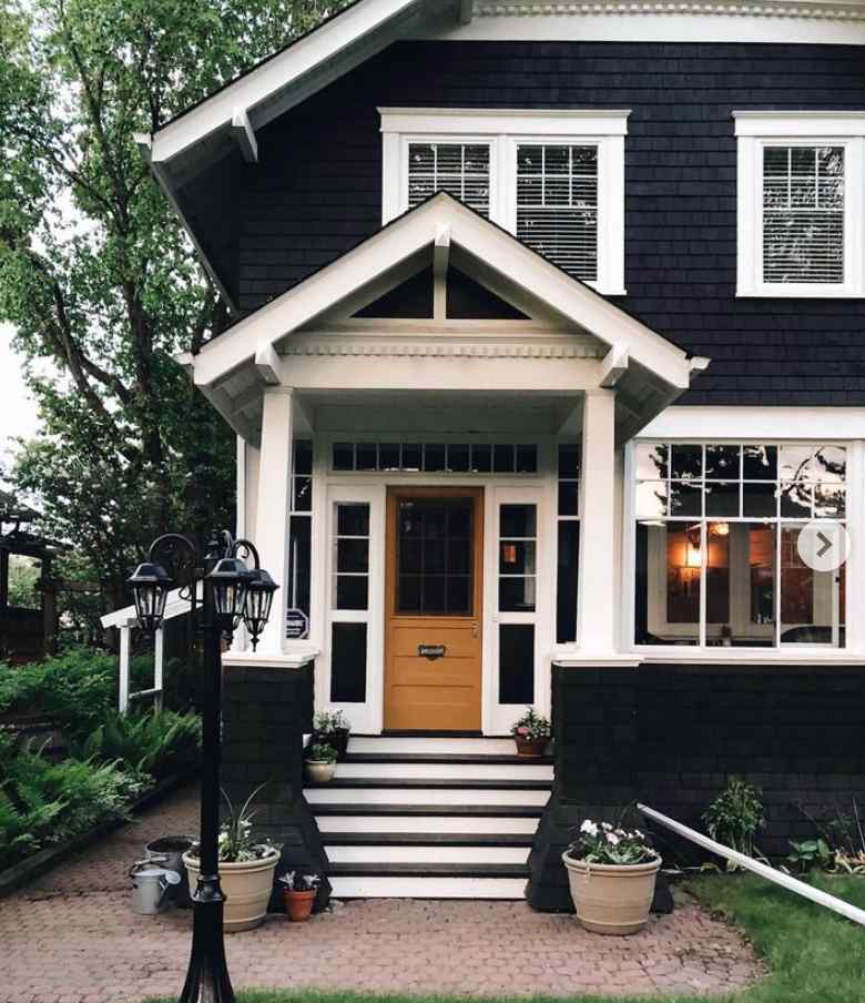 A dark black house with white trim and wooden door and light and a plant pot outside of the house