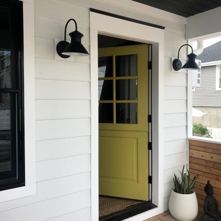 Black barn-style lights on entryway on white house and yellow door and plant pot outside of the door