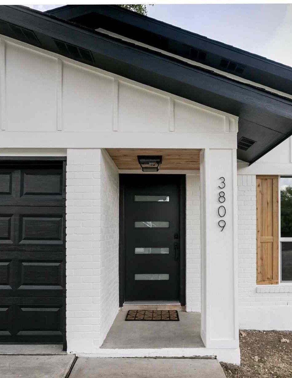 A white house with black house number and black door and mat on the floor and light above the wall