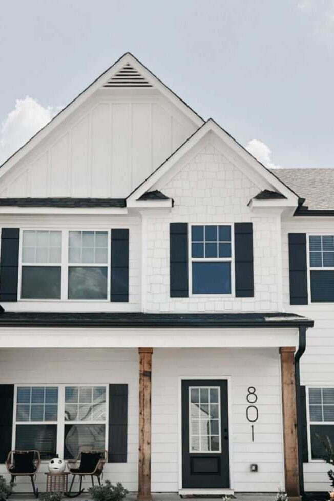 A white house with black house number and black shutter and wooden pillars 