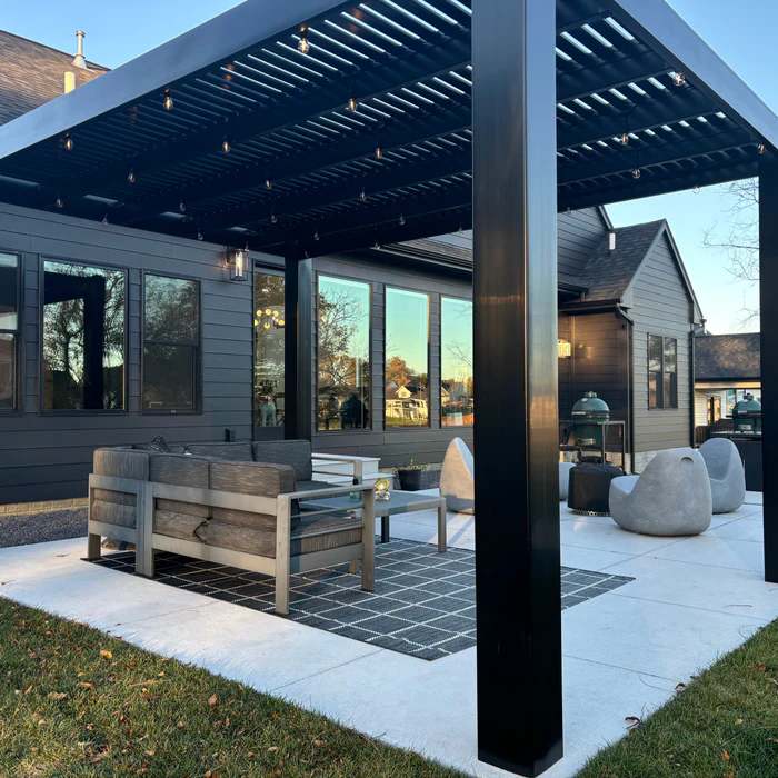 Black pergola with grey house and lights on the wall and black framed windows and sofa outside of the house