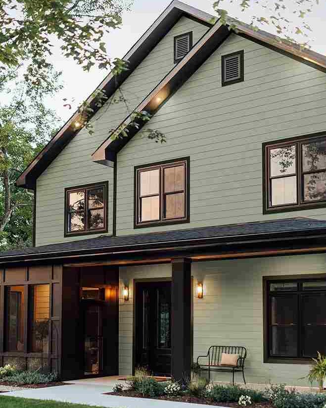 Green house with black trim windows and lights on the wall and chair outside of the house