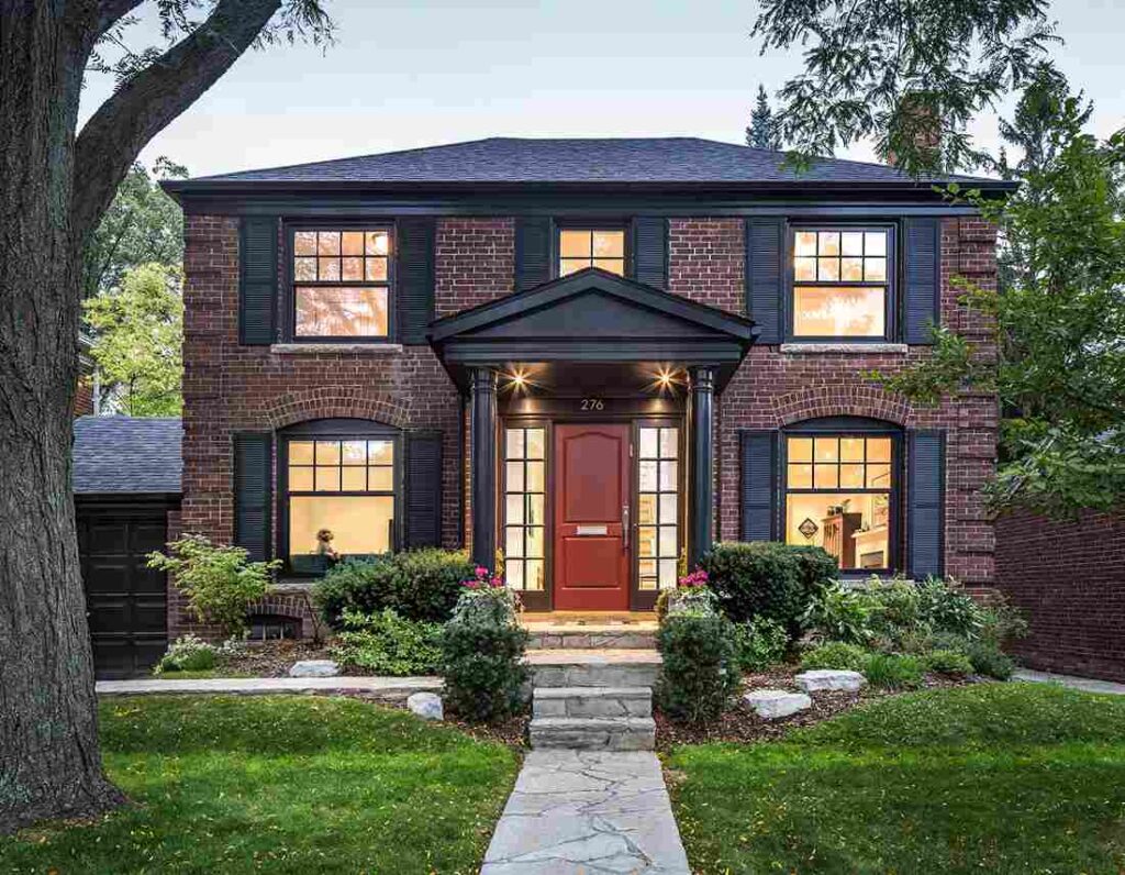 Red brick house with black trim and shutter and glass windows and a pathway and wooden door and celling lights above the front door wall