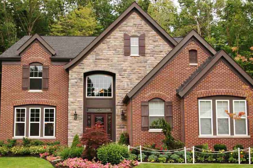 Light tan house and red brick and white trim windows and brown shutter and garden outside of the house