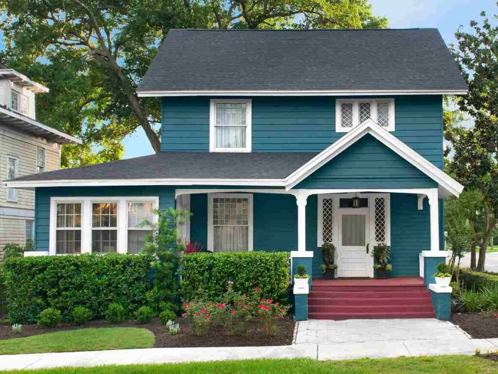 A blue house with green undertones and red stairs and white trim and garden outside of the house
