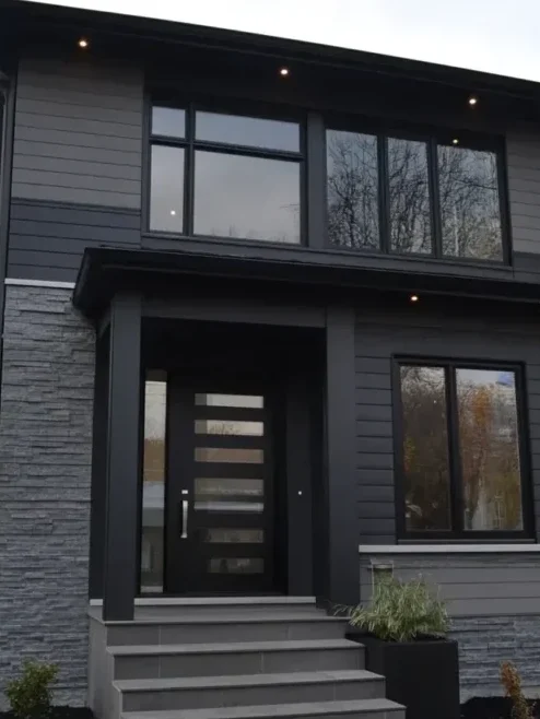 A grey house with black trim and glass windows and lights on the wall
