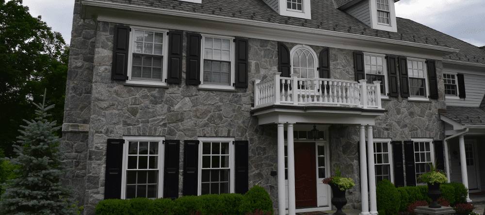 A grey stone house with black shutter and white window and pillars and white balcony