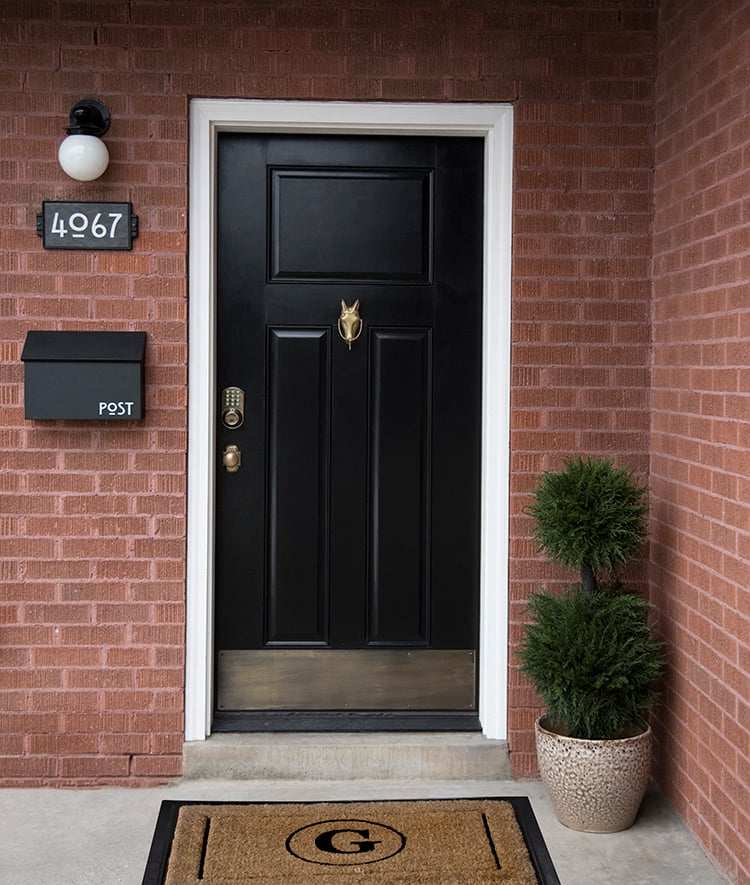A brick house with house number and mat on the floor and plants in the corner 