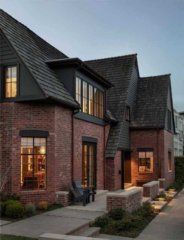 Red brick house with black roof and warm light and chairs outside of the house