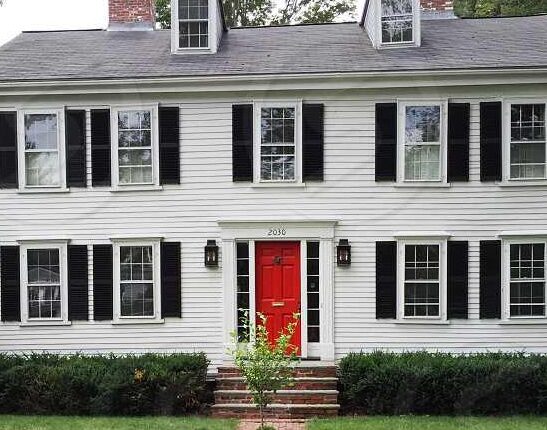 White brick house with red door and black shutter and lights on the wall 