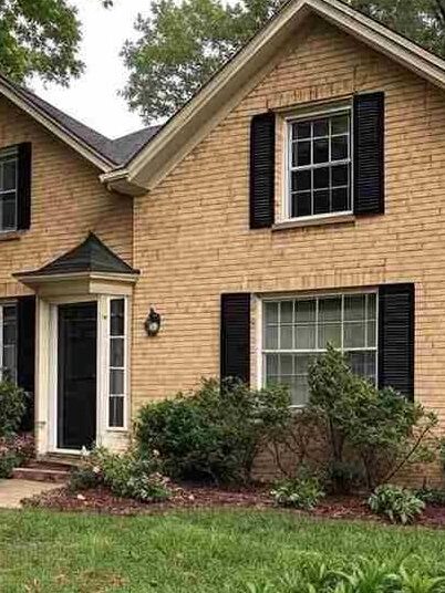 A tan brick house with black shutter and white trim windows and garden outside of the house