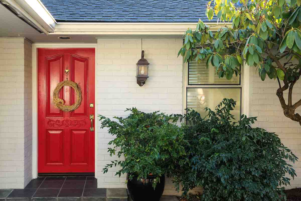 White brick house  with red door and celling light on the wall and plants outside of the house 