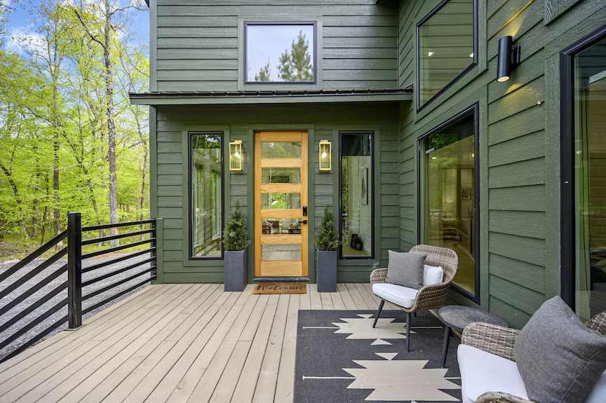 Green house with wooden deck and black trim and sofa chairs outside of the house and glass large windows