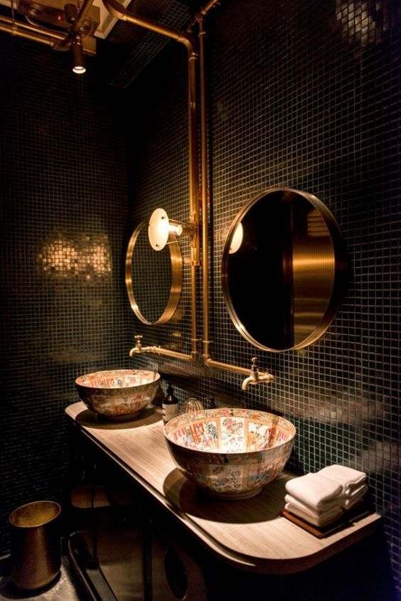 A black bathroom with gold sink and light on wall and round mirror
