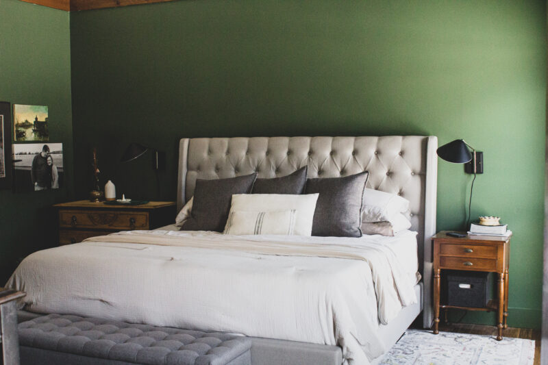 A green bedroom with grey bedding and wooden nightstand and black fixtures 