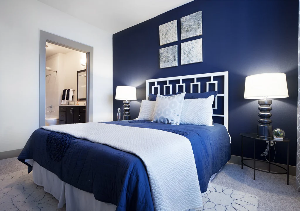 A grey and blue bedroom with white bedding and navy blue blanket and grey rug and grey floor 