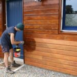 a-person-is-applying-a-finish-likely-a-wood-stain-to-the-exterior-horizontal-siding-of-a-building-and-the-siding-appears-to-be-made-of-western-red-cedar