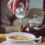 a-white-cup-filled-with-a-tea-and-glass-teapot-with-a-dark-liquid-inside-it-and-hand-is-holding-a-small-container-and-pouring-liquid-into-the-cup