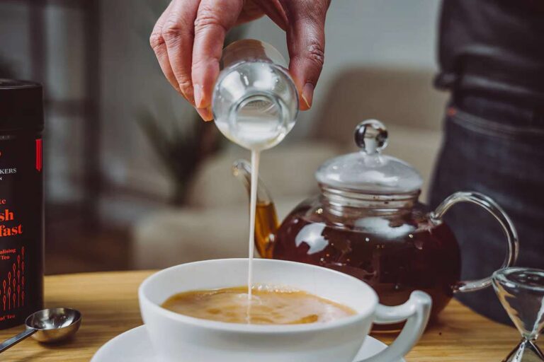 6 Qualities That Define A Balanced Breakfast Tea a-white-cup-filled-with-a-tea-and-glass-teapot-with-a-dark-liquid-inside-it-and-hand-is-holding-a-small-container-and-pouring-liquid-into-the-cup