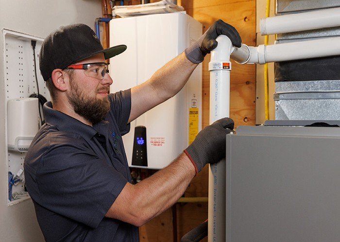 man-wearing-safety-glasses-gloves-and-a-cap-is-adjusting-a-white-vent-pipe-connected-to-a-furnance-unit