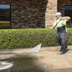 person-wearing-a-white-cap-sunglasses-high-visibilty-vest-over-a-dark-shirt-and-jeans-and-cleaning-ground-with-water