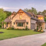 the-house-has-a-multi-level-design-with-mix-of-stone-and-light-colored-siding-on-its-exterior-and-gables-with-decorative-wooden-accents