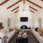 the-room-features-a-high-vaulted-ceiling-with-dark-wood-beams-white-stucco-walls-arched-doorways-and-round-stucco-fireplace-in-the-central-focal-point-of-the-space