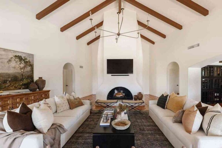 the-room-features-a-high-vaulted-ceiling-with-dark-wood-beams-white-stucco-walls-arched-doorways-and-round-stucco-fireplace-in-the-central-focal-point-of-the-space