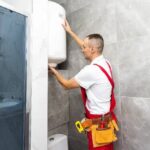 a-man-who-appears-to-be-a-plumber-is-working-inside-a-bathroom-and-standing-beside-a-wall-mounted-water-heater-and-repairing-it