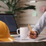 a-person-sitting-at-a-desk-writing-on-papers-a-yellow-construction-hard-hat-is-placed-on-the-desk