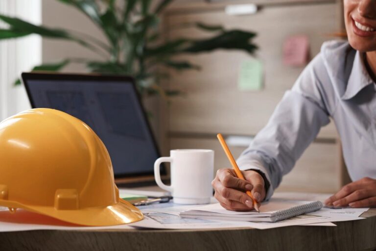 a-person-sitting-at-a-desk-writing-on-papers-a-yellow-construction-hard-hat-is-placed-on-the-desk