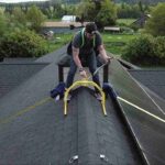 a-person-wearing-a-safety-harness-is-positioned-on-the-peak-of-an-asphalt-shingle-roof