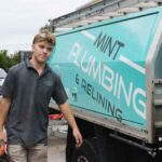 a-plumber-wearing-a-dark-blue-work-shirt-and-light-colored-shorts-is-standing-beside-a-service-truck