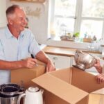 elderly-couple-is-standing-in-a-bright-kitchen-they-are-smiling-and-packing-kitchen-items-into-cardboard-boxes