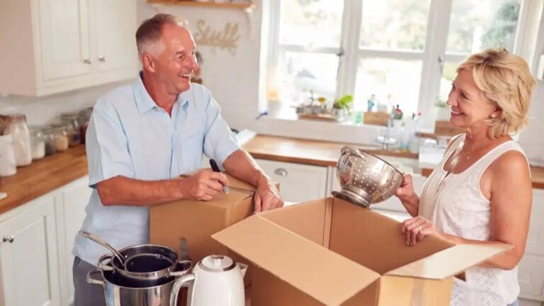 Practical Relocation Tips When Downsizing Your Home elderly-couple-is-standing-in-a-bright-kitchen-they-are-smiling-and-packing-kitchen-items-into-cardboard-boxes