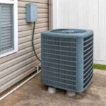 rectangular-blue-gray-metal-air-conditioner-outdoor-unit-sitting-on-the-ground-next-to-the-side-of-a-house