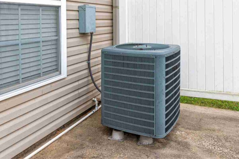 rectangular-blue-gray-metal-air-conditioner-outdoor-unit-sitting-on-the-ground-next-to-the-side-of-a-house