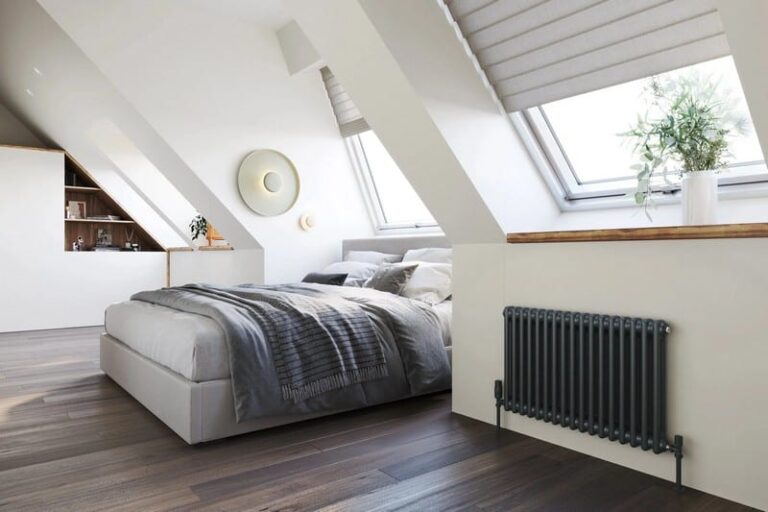 the-room-has-white-sloped-ceilings-and-light-wooden-flooring-and-a-bed-with-white-and-grey-bedding-with-pillows-and-gray-radiator-is-mounted-on-the-wall