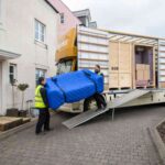 two-movers-carrying-a-large-piece-of-furniture-likely-a-sofa-wrapped-in-a-blue-protective-cover-loading-into-a-yellow-and-white-moving-truck-using-a-metal-ramp