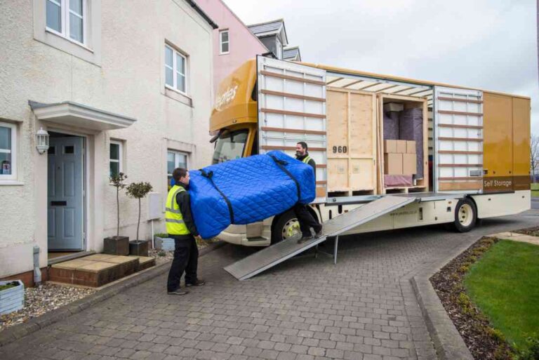How to Minimize Downtime Between Homes When Relocating two-movers-carrying-a-large-piece-of-furniture-likely-a-sofa-wrapped-in-a-blue-protective-cover-loading-into-a-yellow-and-white-moving-truck-using-a-metal-ramp