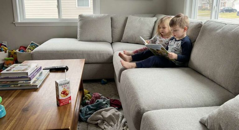 two-young-children-sitting-on-a-light-gray-sectional-sofa-in-a-living-room-and-children-looking-at-a-book-together