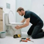 a-man-is-leaning-forward-focused-on-the-base-of-a-white-toilet-appearing-to-be-performing-a-repair-or-installation