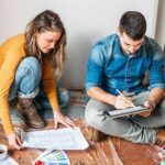 a-woman-in-a-yellow-sweater-and-a-man-in-a-blue-button-shirt-are-looking-at-plans-and-building-material-together