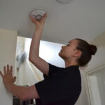 a-woman-reaching-up-to-interact-with-a-white-circular-smoke-alarm-mounted-on-a-ceiling
