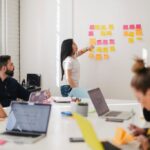 a-woman-stands-at-a-whiteboard-or-wall-covered-in-colorful-sticky-notes-pointing-to-them-while-addressing-a-group-of-colleagues-seated-around-a-table