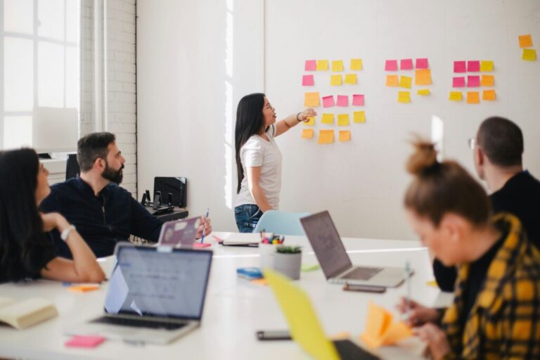 a-woman-stands-at-a-whiteboard-or-wall-covered-in-colorful-sticky-notes-pointing-to-them-while-addressing-a-group-of-colleagues-seated-around-a-table