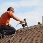 the-roofer-is-kneeling-on-the-shingles-and-using-a-pneumatic-nail-gun-to-secure-the-roofing-material