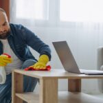 a-man-is-wearing-green-cleaning-gloves-wipedown-or-polishing-a-light-colored-wooden-table-with-a-cloth