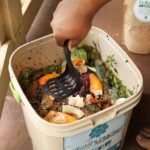 a-person-mixing-food-waste-and-organic-scraps-inside-a-compost-container-using-a-black-scoop