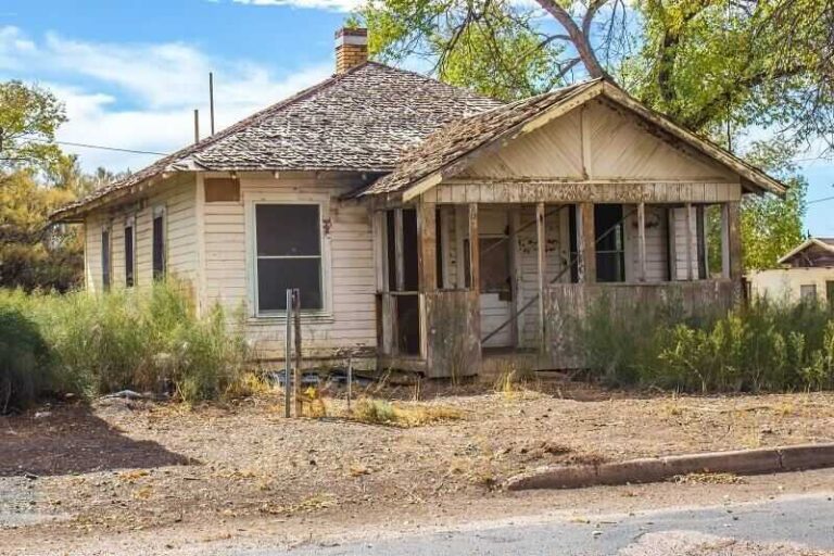 Good Bones, Bad Timing: The Fixer-Upper Problem Smart Buyers Plan For a-single-story-home-features-damaged-front-porch-with-peeling-white-paint-and-a-weathered-wood-shake-roof